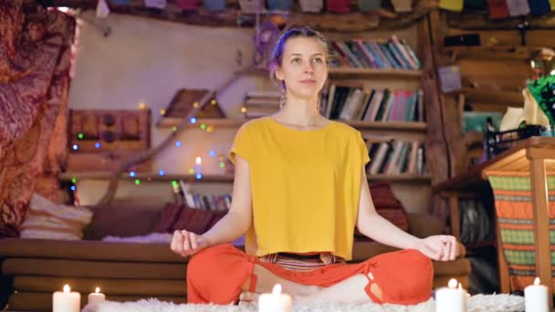 Ein lächelndes schönes junges Mädchen in lockerer Kleidung sitzt in einem Bastelraum auf weißer Fellhaut in einer Lotusposition, umgeben von Kerzen. spirituelles Wachstum und Pflege der eigenen Gesundheit — Stockvideo