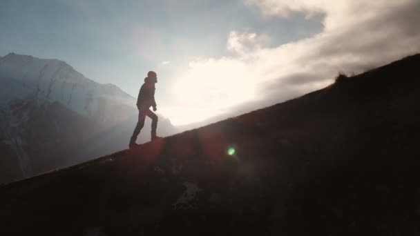 Very long shot Aerial view of epic shot of a man walking on the edge of the mountain as a silhouette in a beautiful sunset. Silhouette of a man with a beard and wearing a hat climbing uphill — Stockvideo