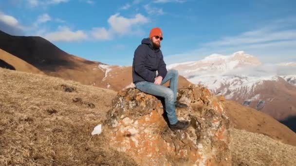 サングラスでひげを生やしたヒップスターは、雪をかぶった山々を背景に、丘の中腹にある大きな石の上に座っている。旅行ビデオ空中ビュー — ストック動画