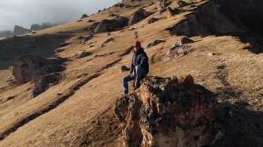 Güneş gözlüğü bir sakallı hipster karla kaplı dağların arka planına karşı bir yamaçta büyük bir taş üzerinde oturuyor. Seyahat videosu havadan görünüm