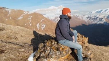 Güneş gözlüğü bir sakallı hipster karla kaplı dağların arka planına karşı bir yamaçta büyük bir taş üzerinde oturuyor. Seyahat videosu havadan görünüm