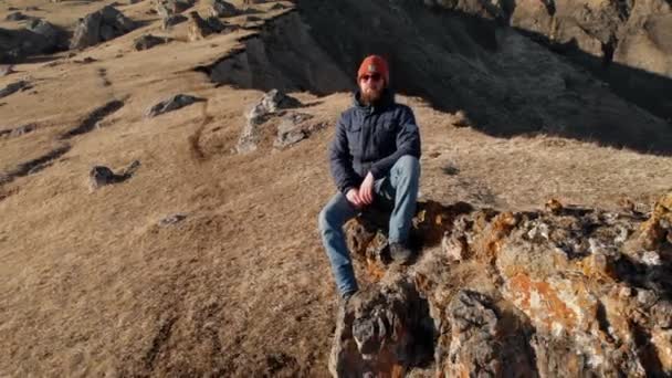 Un hipster barbudo con gafas de sol está sentado en una gran piedra en una ladera sobre el fondo de montañas cubiertas de nieve. Vista aérea de vídeo de viaje — Vídeo de stock