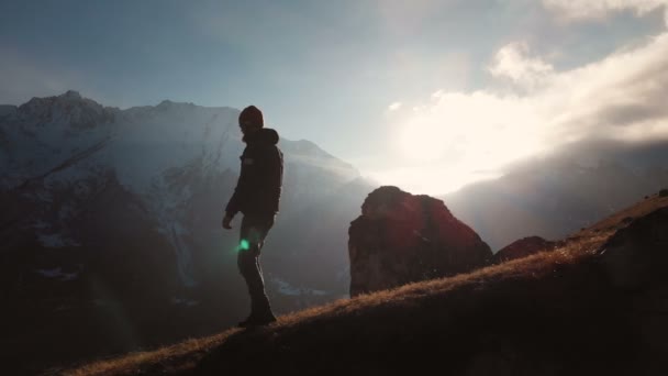 Vista aérea de um tiro épico de um homem caminhando na borda de uma montanha como uma silhueta em um belo pôr do sol. Silhueta de um homem com uma barba e um chapéu descendo a montanha em uma montanha — Vídeo de Stock
