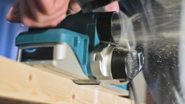 Büyük yakın çekim. Bir marangoz, elektrikli düzlemde ahşap bir tahta kesiyor. Toz parçacıklarının ve testere bıçağının yavaş hareketi