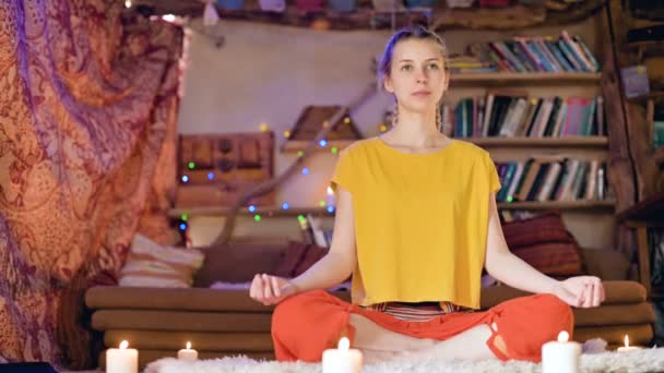 Una joven hermosa y sonriente vestida con ropa suelta se sienta en una sala de manualidades sobre una piel de piel blanca en posición de loto rodeada de velas. Meditación crecimiento espiritual y cuidado de su propia salud — Vídeos de Stock