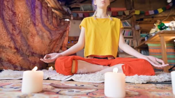 A beautiful young girl in loose clothes sits in a craft room on a white fur skin in a lotus position surrounded by candles. Meditation spiritual growth and care for your own health — Stock Video