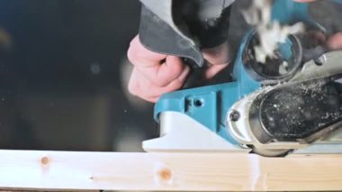 Büyük yakın çekim. Bir marangoz, elektrikli düzlemde ahşap bir tahta kesiyor. Toz parçacıklarının ve testere bıçağının yavaş hareketi