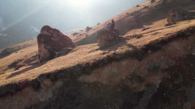 Güneş gözlüğü bir sakallı hipster karla kaplı dağların arka planına karşı bir yamaçta büyük bir taş üzerinde oturuyor. Seyahat videosu havadan görünüm