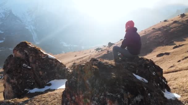 Um hipster barbudo em óculos de sol está sentado em uma grande pedra em uma encosta no fundo de montanhas cobertas de neve. Vídeo de viagem vista aérea — Vídeo de Stock