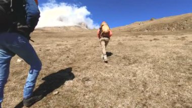 Şapka ve güneş gözlüğü sırt çantaları ile iki turist fotoğrafçılar karlı dağların arka planına karşı ellerinde kameralar ile sarı çim tepede yukarı gidin. yavaş hareket