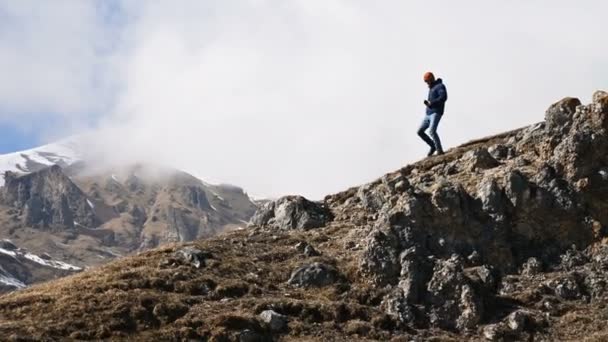 Ο μουσάτος νέος άντρας φωτογράφος με καπέλο και γυαλιά ηλίου και μια κάμερα γύρω από το λαιμό του σκαρφαλώνουν στο λόφο με φόντο τα χιονισμένα βουνά και τα σύννεφα — Αρχείο Βίντεο