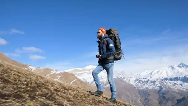 Бородатый молодой фотограф в шляпе и солнцезащитных очках с большим рюкзаком и камерой вокруг шеи взбирается на холм на фоне заснеженных гор — стоковое видео
