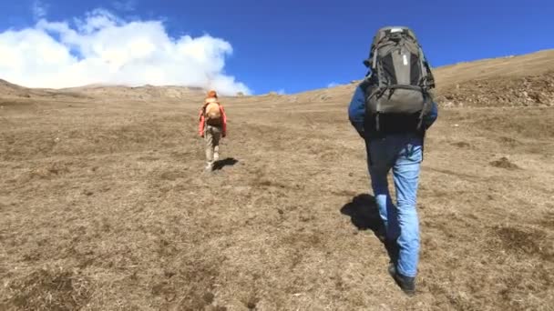 Dos fotógrafos turistas con mochilas en sombreros y gafas de sol suben la colina en la hierba amarilla con cámaras en sus manos contra el fondo de las montañas nevadas. cámara lenta — Vídeos de Stock