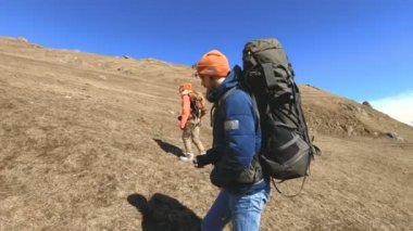 Şapka ve güneş gözlüğü sırt çantaları ile iki turist fotoğrafçılar karlı dağların arka planına karşı ellerinde kameralar ile sarı çim tepede yukarı gidin. yavaş hareket