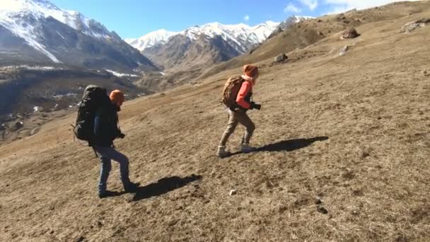 Två turister fotografer med rygg säckar i hattar och sol glasögon gå upp för backen på det gula gräset med kameror i sina händer mot bakgrund av snöiga berg. långsam rörelse — Stockvideo