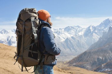 Güneş gözlüğü ve güneşli bir günde karlı dağların arka planına karşı sırtında bir sırt çantası ile sıcak bir ceket bir sakallı adam portresi. Turizm ve seyahat konsepti