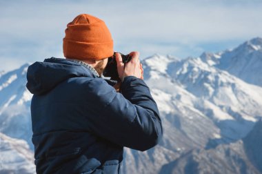 Güneş gözlüğü bir sakallı erkek fotoğrafçı ve sırtında bir sırt çantası ile sıcak bir ceket ve elinde bir refleks kamera portresi karla kaplı dağların arka planına karşı fotoğraf çeker