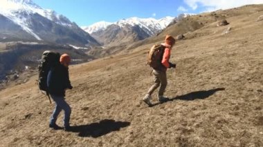 Şapka ve güneş gözlüğü sırt çantaları ile iki turist fotoğrafçılar karlı dağların arka planına karşı ellerinde kameralar ile sarı çim tepede yukarı gidin. yavaş hareket