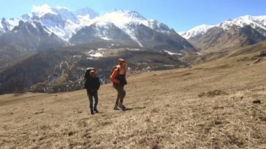 Şapka ve güneş gözlüğü sırt çantaları ile iki turist fotoğrafçılar karlı dağların arka planına karşı ellerinde kameralar ile sarı çim tepede yukarı gidin. yavaş hareket