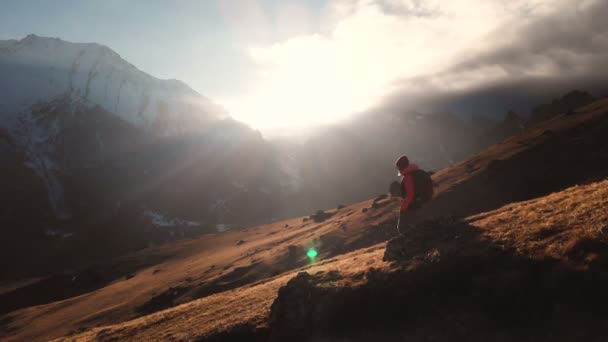 Vista aérea de una foto épica de una niña caminando en el borde de una montaña como una silueta en una hermosa puesta de sol. Silueta de una chica en un sombrero con una mochila bajando por la montaña en una ladera de la montaña — Vídeo de stock
