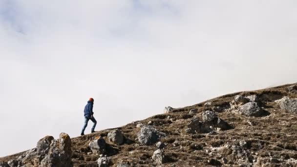 Joven fotógrafo barbudo con sombrero y gafas de sol y una cámara alrededor de su cuello sube la colina sobre el fondo de montañas nevadas y nubes — Vídeo de stock