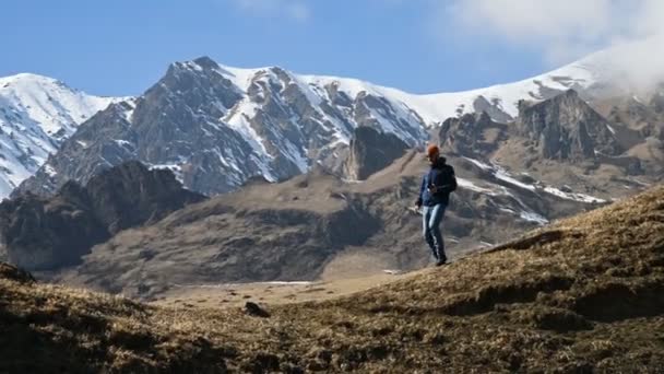 Skäggiga unga manliga fotograf i en hatt och sol glasögon och en kamera runt halsen klättrar upp för backen mot bakgrund av snötäckta berg och moln — Stockvideo