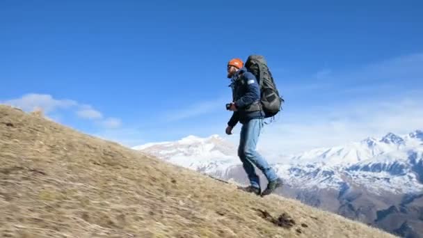 Büyük bir sırt çantası ve boynunun etrafında bir kamera ile bir şapka ve güneş gözlüğü sakallı genç erkek fotoğrafçı karla kaplı dağların arka planına karşı tepeye tırmanıyor — Stok video