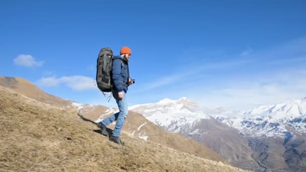 Bärtige junge männliche Fotograf mit Hut und Sonnenbrille mit einem großen Rucksack und einer Kamera um den Hals kommt den Hügel hinunter von einem Berg vor der Kulisse der schneebedeckten Berge — Stockvideo
