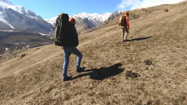 Två turister fotografer med rygg säckar i hattar och sol glasögon gå upp för backen på det gula gräset med kameror i sina händer mot bakgrund av snöiga berg. långsam rörelse — Stockvideo