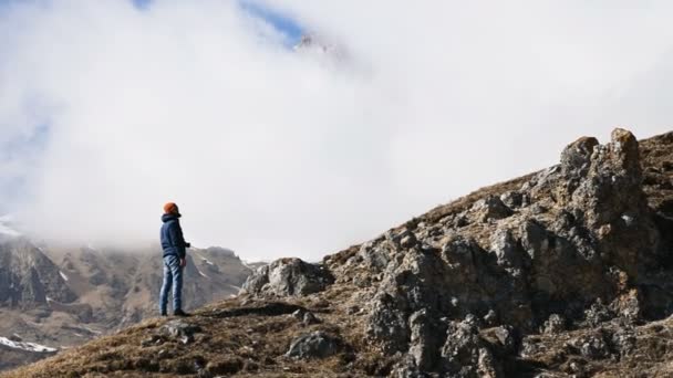 Joven fotógrafo barbudo con sombrero y gafas de sol y una cámara alrededor de su cuello sube la colina sobre el fondo de montañas nevadas y nubes — Vídeos de Stock