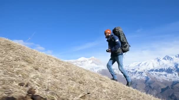 Jovem fotógrafo barbudo em um chapéu e óculos de sol com uma grande mochila e uma câmera ao redor de seu pescoço sobe a colina contra o fundo de montanhas cobertas de neve — Vídeo de Stock