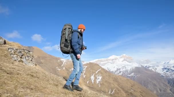 Giovane fotografo maschio barbuto con cappello e occhiali da sole con un grande zaino e una macchina fotografica intorno al collo che scende dalla collina da una montagna sullo sfondo di montagne innevate — Video Stock