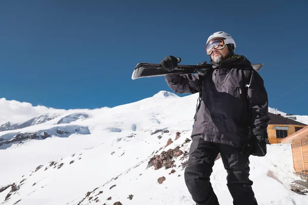 Ritratto barbuto sciatore maschile invecchiato sullo sfondo di montagne innevate del Caucaso. Un uomo adulto che indossa maschera da sci googles e sci casco sulla spalla guarda montagne. Concetto di stazione sciistica — Foto Stock