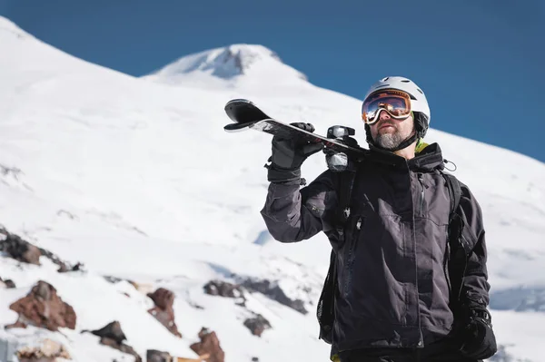 Esquiador macho barbudo retratado envelhecido no fundo de montanhas de Cáucaso cobertas de neve. Um homem adulto usando óculos de esqui máscara e esquis capacete em seu ombro parece montanhas. Conceito de resort de esqui — Fotografia de Stock