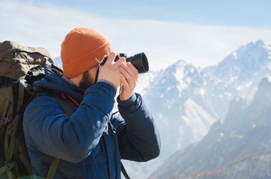 Güneş gözlüğü bir sakallı erkek fotoğrafçı ve sırtında bir sırt çantası ile sıcak bir ceket ve elinde bir refleks kamera portresi karla kaplı dağların arka planına karşı fotoğraf çeker