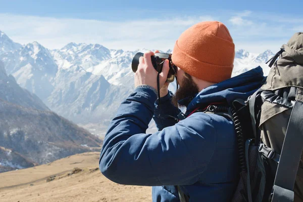Güneş gözlüğü bir sakallı erkek fotoğrafçı ve sırtında bir sırt çantası ile sıcak bir ceket ve elinde bir refleks kamera portresi karla kaplı dağların arka planına karşı fotoğraf çeker