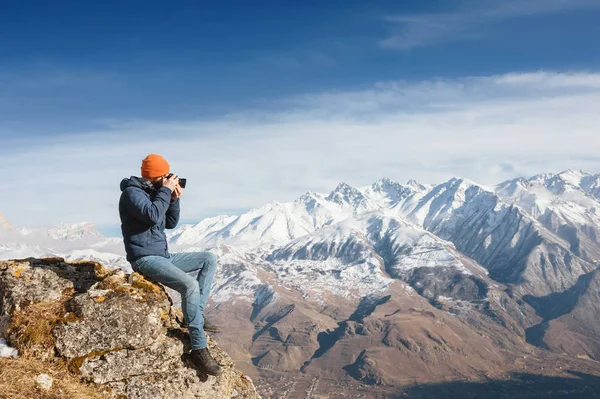 Portrait of a bearded traveler photographer in sunglasses and a cap sits on a rock with mirror camera in his hands against the backdrop of mountains