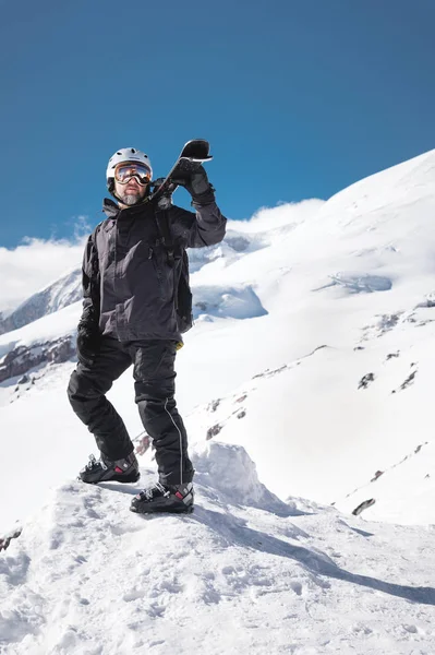Retrato de Crescimento Esquiador macho barbudo envelhecido contra o fundo das montanhas do Cáucaso cobertas de neve. Um homem adulto usando óculos de esqui máscara e esquis capacete em seu ombro parece montanhas. Conceito de resort de esqui — Fotografia de Stock
