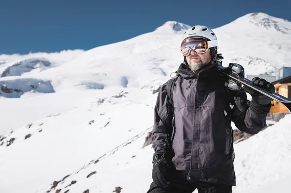 Esquiador macho barbudo retratado envelhecido no fundo de montanhas de Cáucaso cobertas de neve. Um homem adulto usando óculos de esqui máscara e esquis capacete em seu ombro parece montanhas. Conceito de resort de esqui — Fotografia de Stock