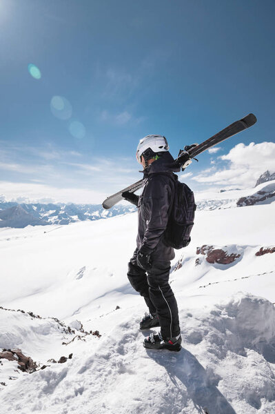 Growth Portrait bearded male skier aged against background of snow-capped Caucasus mountains. An adult man wearing ski googles mask and helmet skis on his shoulder looks mountains. Ski resort concept