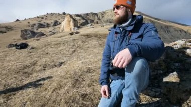 Güneş gözlüğü ve bir kap sakallı bir gezgin fotoğrafçı portresi dağların arka planında elinde ayna kamera ile bir kaya üzerinde oturur