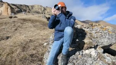 Güneş gözlüğü ve bir kap sakallı bir gezgin fotoğrafçı portresi elinde ayna kamera ile bir kaya üzerinde oturur ve dağların arka planında bir resim çekmek