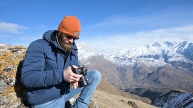 Güneş gözlüğü ve bir kap sakallı bir gezgin fotoğrafçı portresi elinde ayna kamera ile bir kaya üzerinde oturur ve dağların arka planında bir resim çekmek