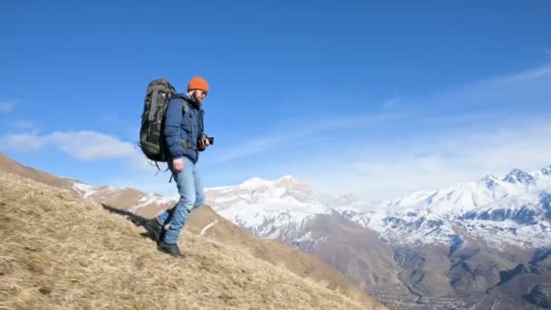 Bärtige junge männliche Fotograf mit Hut und Sonnenbrille mit einem großen Rucksack und einer Kamera um den Hals kommt den Hügel hinunter von einem Berg vor der Kulisse der schneebedeckten Berge — Stockvideo