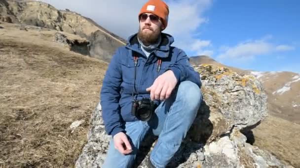 Retrato de um viajante barbudo fotógrafo em óculos de sol e um boné senta-se em uma rocha com câmera de espelho em suas mãos contra o pano de fundo das montanhas — Vídeo de Stock