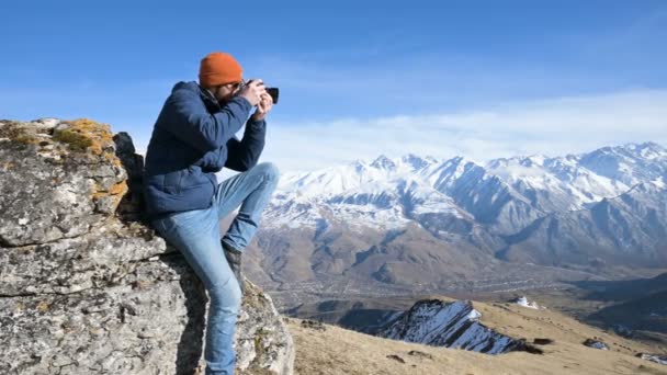 Güneş gözlüğü ve bir kap sakallı bir gezgin fotoğrafçı portresi elinde ayna kamera ile bir kaya üzerinde oturur ve dağların arka planında bir resim çekmek — Stok video