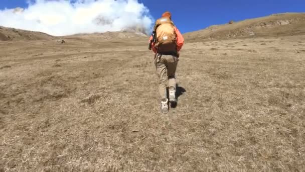 Feliz fotógrafa viajera en gafas de sol con una cámara y una mochila va cuesta arriba sobre un fondo de picos nevados en un día soleado — Vídeos de Stock