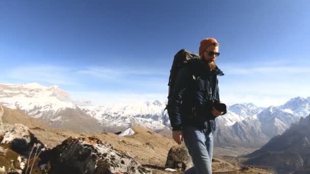 Buon viaggiatore uomo barbuto fotografo in occhiali da sole con una macchina fotografica e uno zaino va in salita su uno sfondo di cime innevate in una giornata di sole — Video Stock