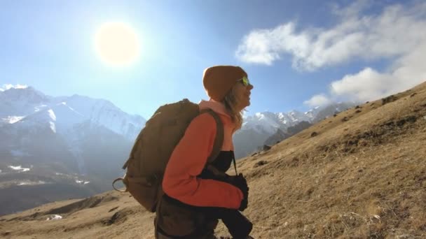 Glückliche Reisende Fotografin in Sonnenbrille mit Kamera und Rucksack geht bergauf auf einem Hintergrund von schneebedeckten Gipfeln an einem sonnigen Tag — Stockvideo