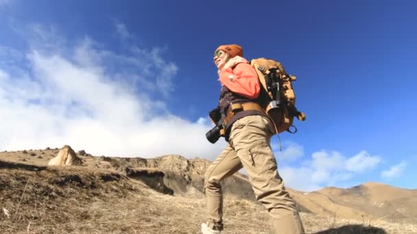 Šťastná fotografka z cestičky v brýlích s kamerou a batohu vede do kopce na pozadí zasněžené špičky za slunečného dne — Stock video